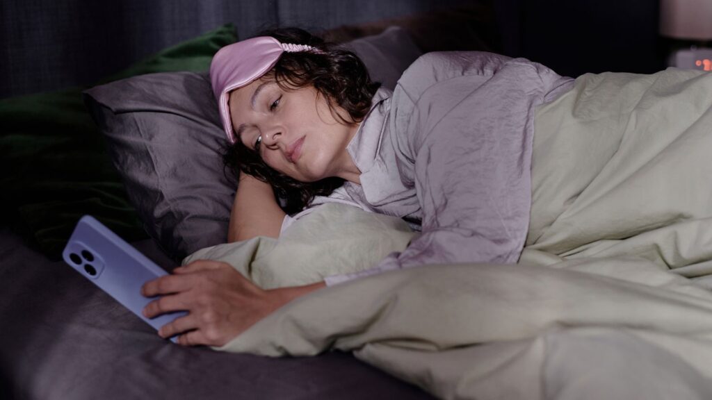 Kobieta w piżamie leży w łóżku, przeglądając pocztę na telefonie przed snem.