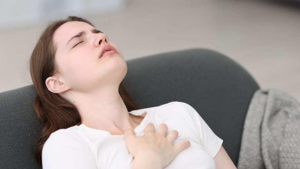 Kobieta leży na kanapie z ręka na klatce piersiowej i z zamkniętymi oczami, próbuje zredukować stres.