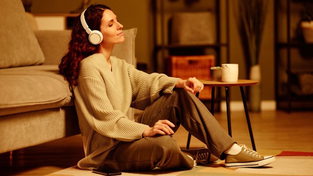 Kobieta siedzi oparta o sofę z słuchawkami na uszach i relaksuje się słuchając muzyki.