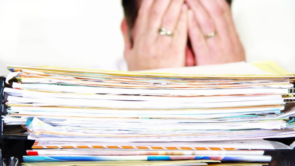 Zrezygnowany i zmęczony mężczyzna kładzie ręce na twarz mając przed sobą stos dokumentów.