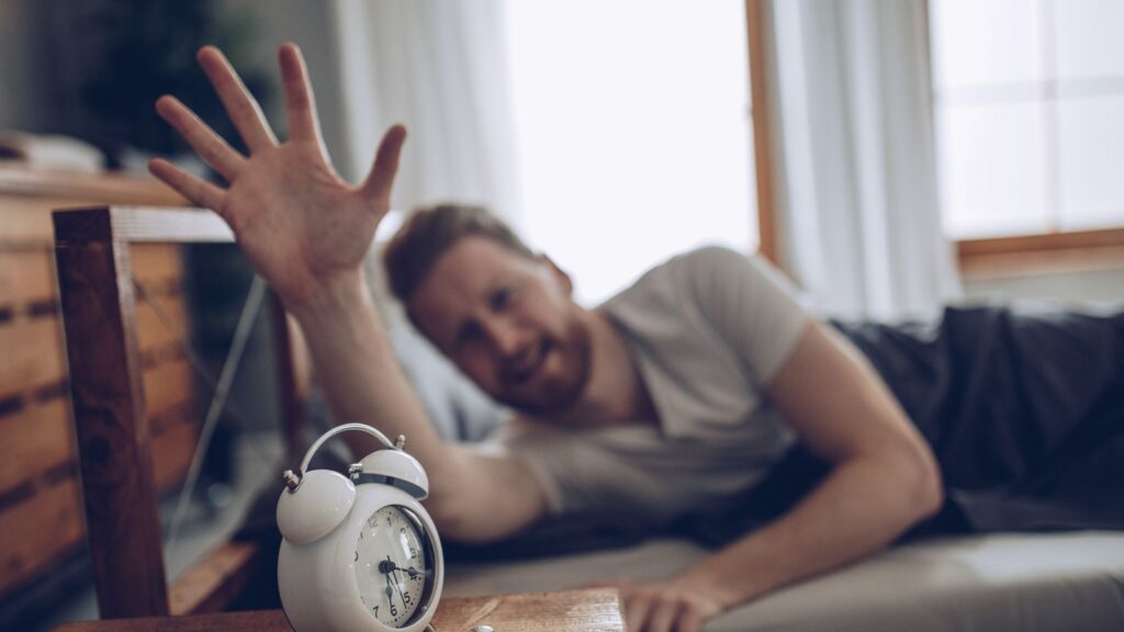 Mężczyzna budzi się rano i próbuje wyłączyć budzik, bo jest zmęczony.