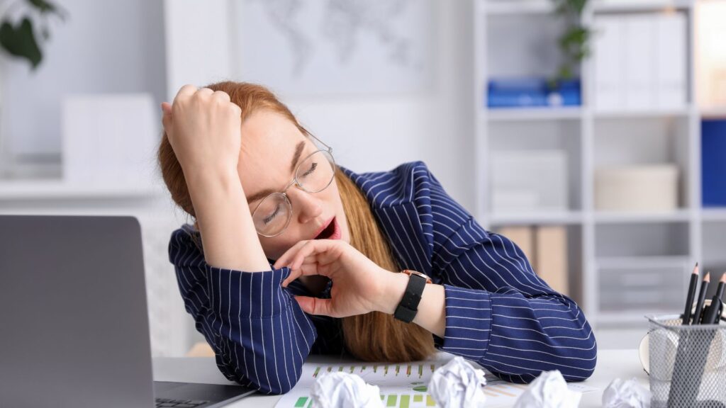 Zmęczona kobieta siedzi przy biurku i ziewa, dając wyraz zmęczeniu po wielu godzinach pracy.
