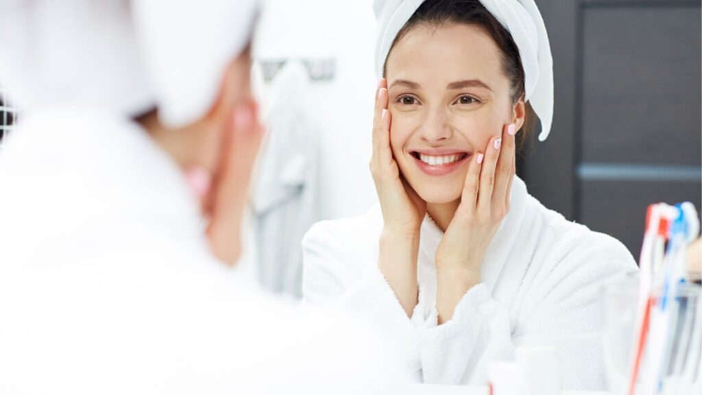 Kobieta z ręcznikiem na głowie patrzy w lustro podczas porannej rutyny pielęgnacyjnej.